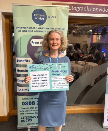 A smiling woman holds up a signed Employer's Pledge poster in front of a display stand advertising Citizens Advice Bucks.