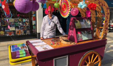 A man smiling and wearing a bright pink wig standing behind at purple cart in Bracknell Town Centre. The cart is purple and has lots of colourful paper decorations. On the cart are a variety of leaflets about Buckinghamshire Mind's services.