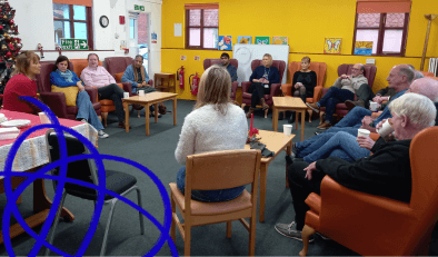 A groups of people sitting in chairs, mostly armchairs, in a circle in a room. There are small tables with cups of tea or coffee on them.