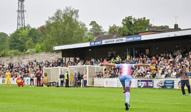 A Chesham United football players running across the pitch with his head down and his arms outstretched. Shown from the back. There is a crowd cheering in the stands in front of him and a photographer taking a photo.