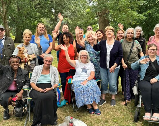 A diverse group of people attending a Friends in Need picnic. They are all smiling and waving their hands.
