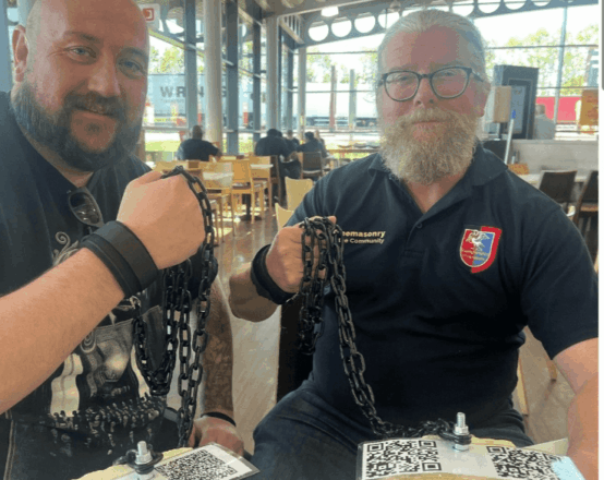 Ewan Gordon standing with another man (on his left). Both are smiling and holding up the chains that link each of them to the Rough Ashlar.