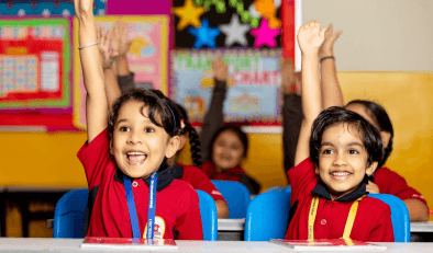 A diverse group of school children sitting at desk with their hands up and smiling.