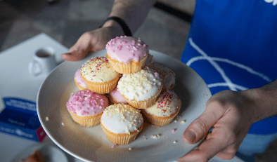 A plate of cakes being held up. The hands of the person holding it are visible In the background a Bucks Mind collection box is visible.