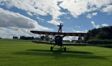 The sillouhette of a person standing on top of the wings of a bi-plane in a field. There is blue sky and white clouds behind the person and plan.