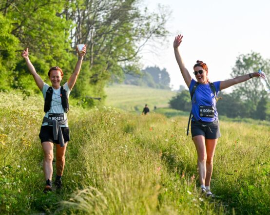 Two female competitors smiling with their arms in the air in a field. Another competitor is in the distant background.